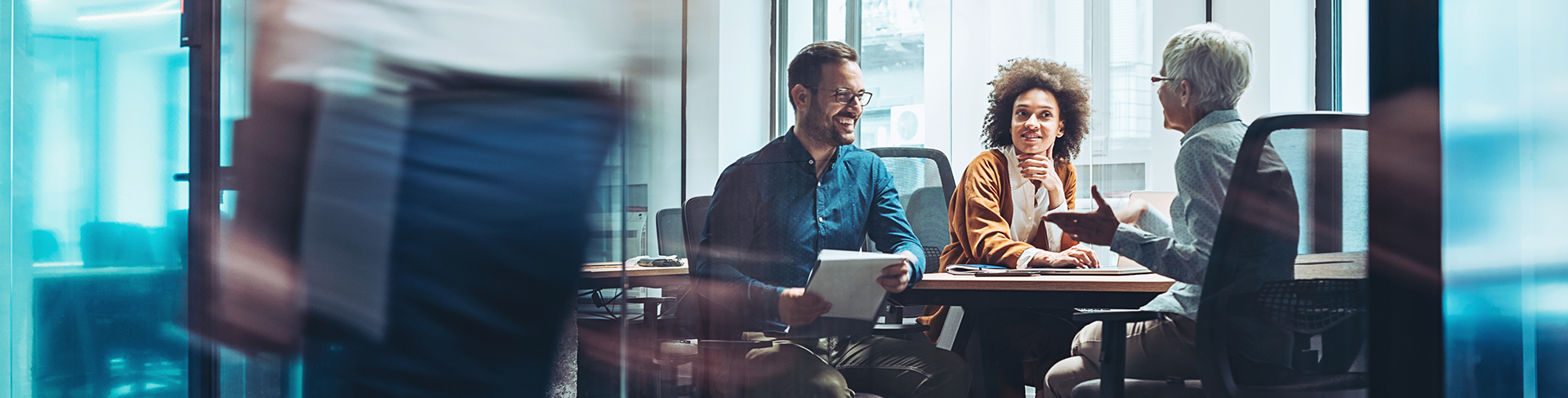 Three colleagues having a positive meeting in a glass-walled office, while another person walks by in a motion blur.