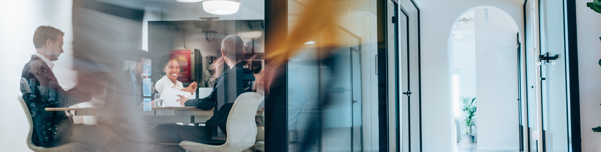 A busy modern office hallway with motion-blurred people walking past a glass-walled conference room where a meeting is in progress.