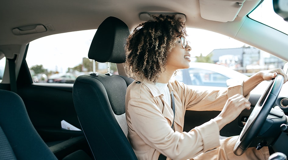 A woman in glasses and a coat drives a car, as she asks herself whether she can let someone else drive her car