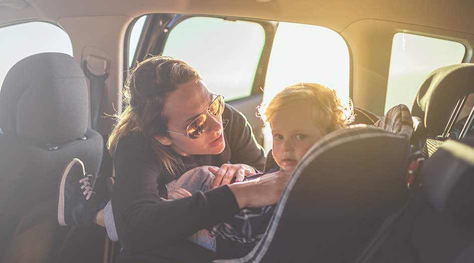 A woman buckles her young child into a car seat in the back of a vehicle, reducing risk of injury if airbags were to deploy.