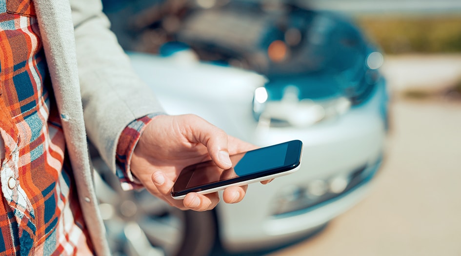 A man holds his smart phone with a car in the background, showing some simple ways to avoid the most common car insurance claims