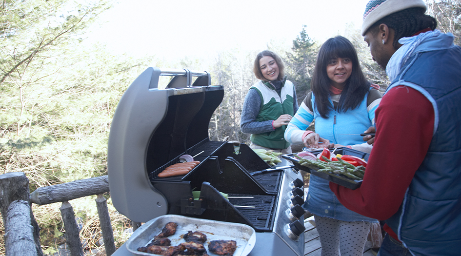 Three individuals wear jackets and add food to a BBQ, showing you how to engage in safe winter grilling