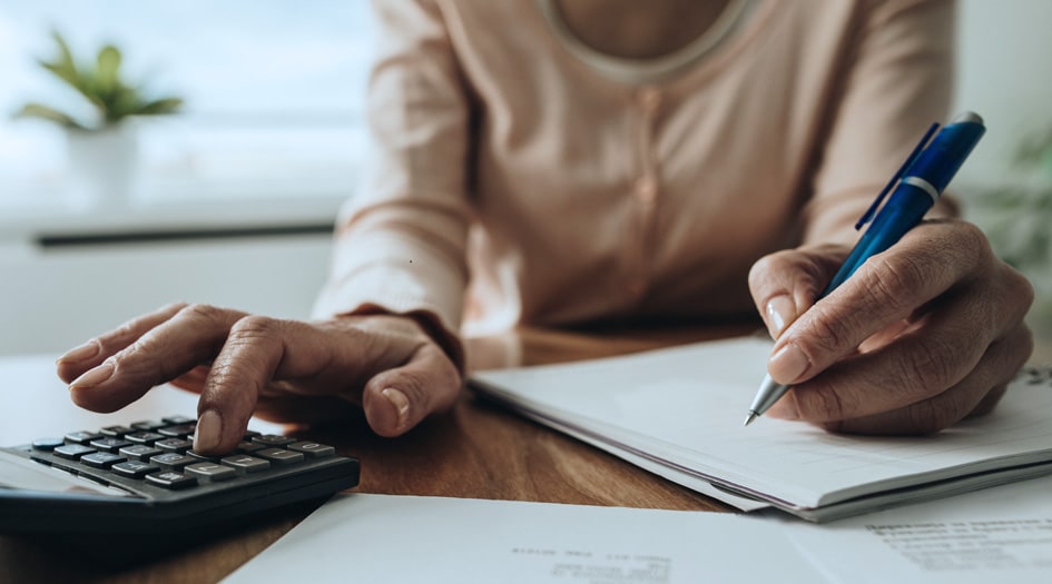 A close-up shows an individual writing on a piece of paper and typing on a calculator, as they save money for a house.