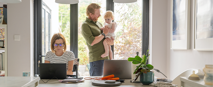 Une femme travaille sur son ordinateur portable à une table, tandis qu'un homme tient leur enfant en arrière-plan.
