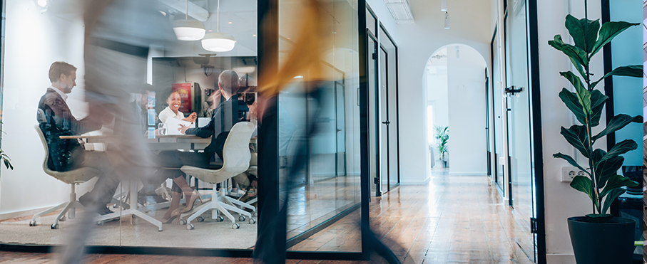 A busy modern office hallway with motion-blurred people walking past a glass-walled conference room where a meeting is in progress.