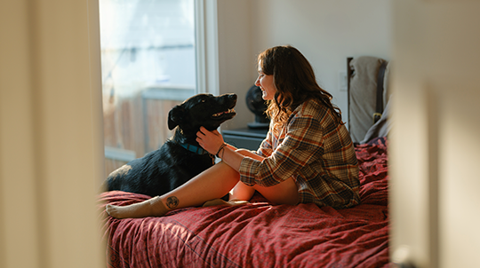 Une jeune femme vêtue d'une chemise à carreaux est assise sur un lit aux draps rouges, souriant en caressant son chien noir sous le menton. La pièce est baignée d'une lumière naturelle et chaude provenant d'une fenêtre.