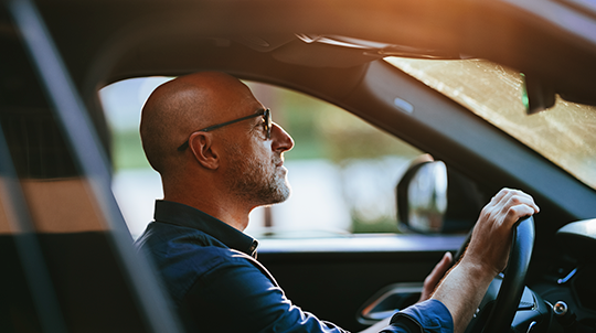 Vue de profil d'un homme chauve à lunettes, concentré au volant de sa voiture, le visage illuminé par la lumière chaude du soleil couchant ou levant.
