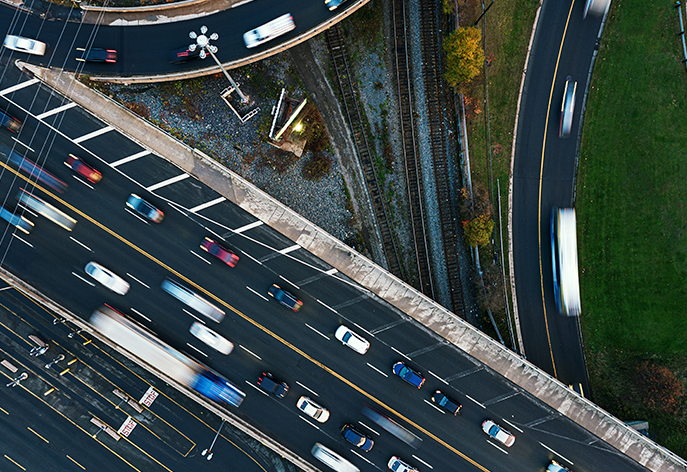 Une photographie aérienne en plongée d'un échangeur autoroutier achalandé où plusieurs routes et voies ferrées se croisent. La longue exposition crée un flou de mouvement, montrant le flux constant de la circulation.