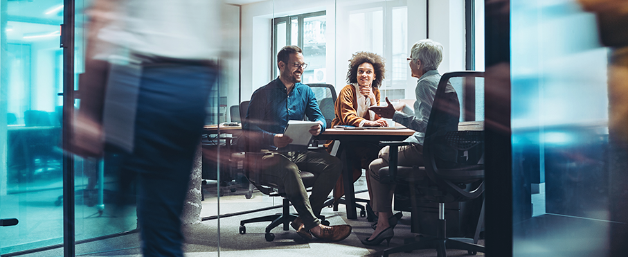 Three colleagues having a positive meeting in a glass-walled office, while another person walks by in a motion blur.
