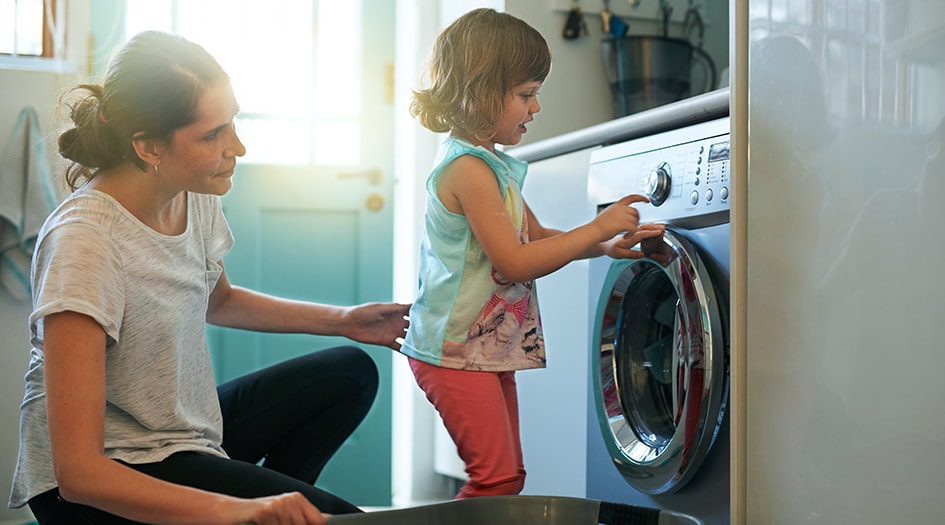 Une femme surveille son enfant en appuyant sur les boutons de la machine à laver, vous montrant comment prévenir les feux de sécheuse.  