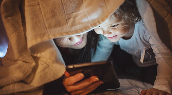 Blottis sous une couverture, une femme et son enfant font des recherches en ligne sur ce que vous devez faire avant, pendant et après une panne d’électricité en hiver.