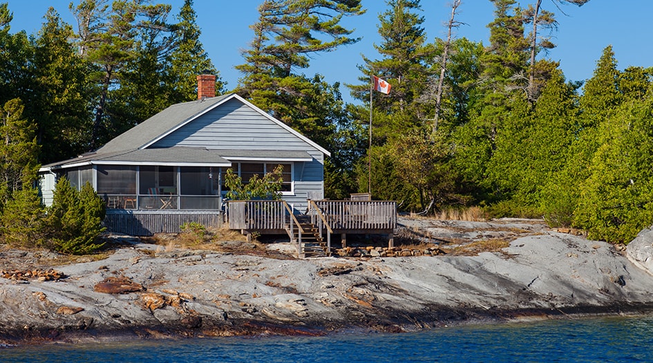 Un petit chalet blanc repose sur la rive d’un lac, pour illustrer comment préparer votre chalet pour l’été.